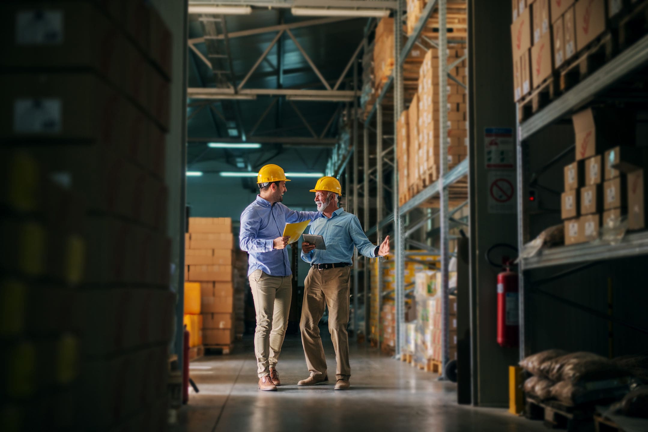 Two men in hard hats smiling and conversing in a warehouse