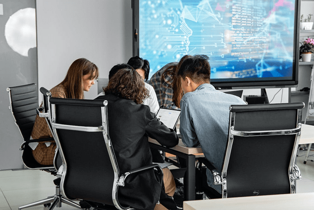 A small group of seated people lean in together around a desk with a large screen displaying data in the background.