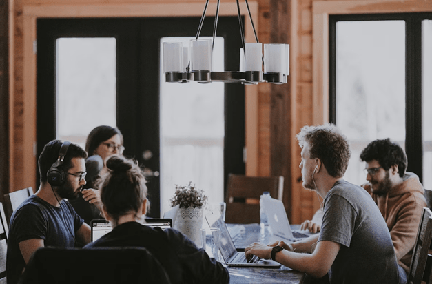 People meeting together sit around a table and take notes on laptops.