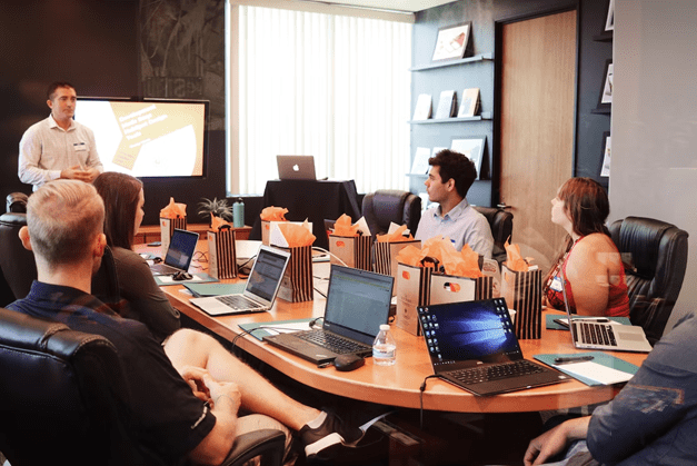 Man presenting to a room of people with laptops, seated around a large table.