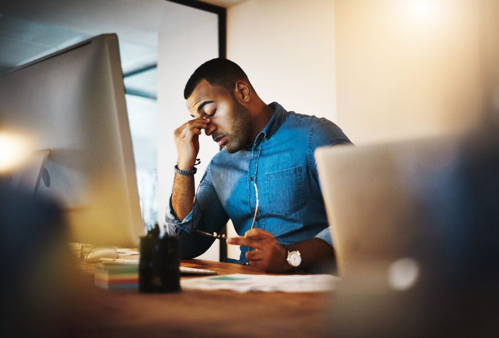 A man sits behind his computer, looking stressed as he rubs his face with one hand.