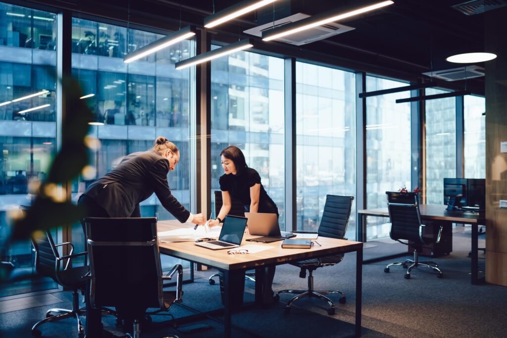 Two people are standing across a table in an office, discussing papers