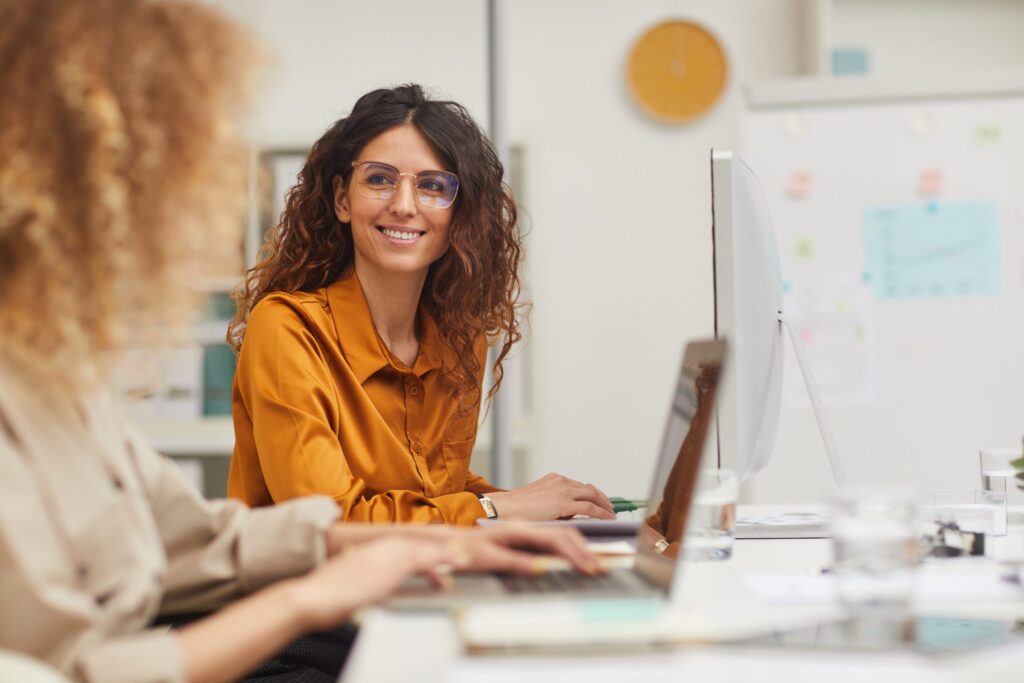 Two women are sitting at desks in an office, each working on a device while engaged in a conversation