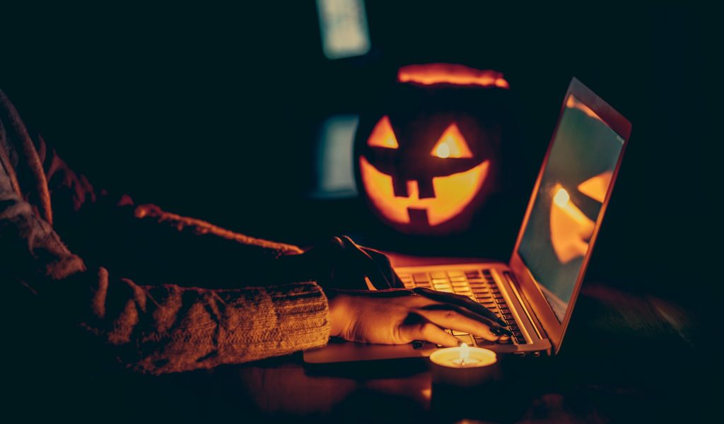 The image features a Halloween-themed scene with a woman's hand typing on a laptop. In the foreground, there's a lit candle, while in the background, a carved pumpkin with a candle inside