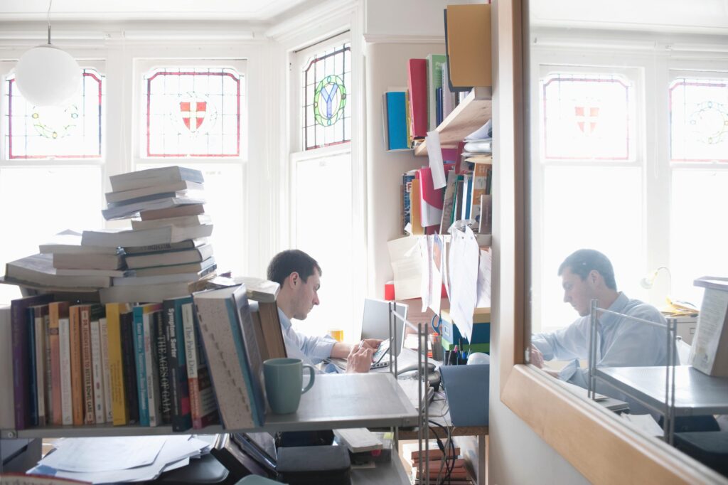 In the foreground, there is a stack of books, while in the background, a man is working on a laptop, with his reflection visible in a nearby mirror.