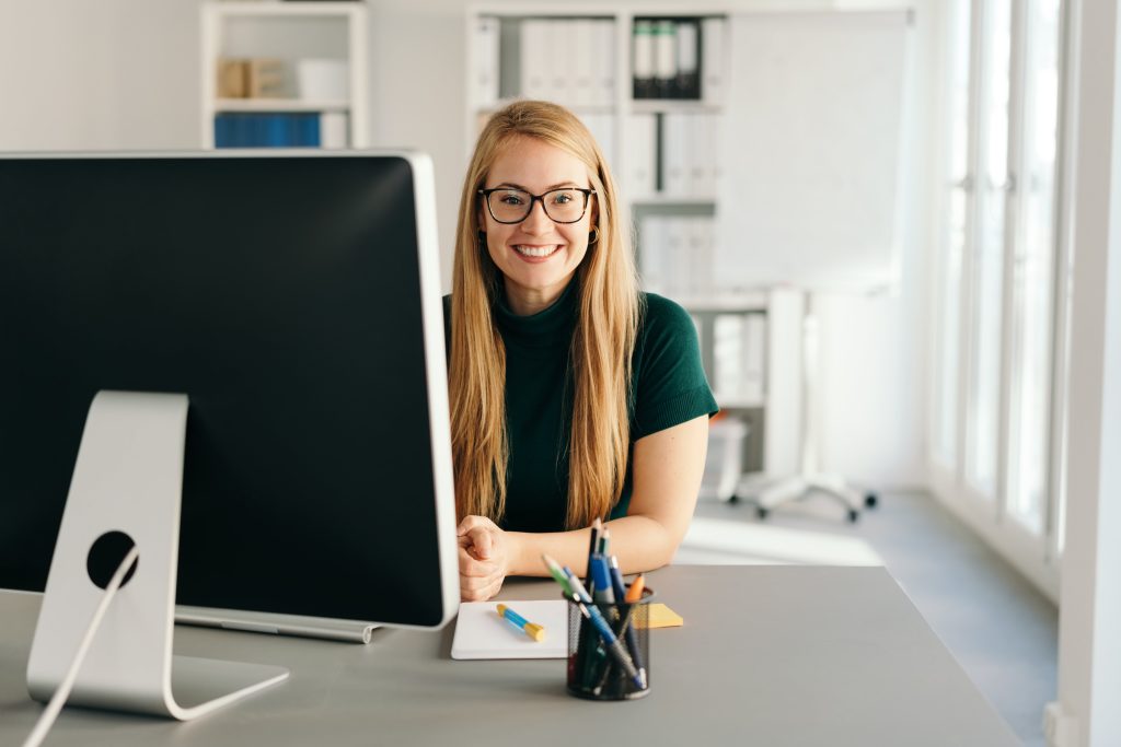 A woman is sitting in front of her desktop in an office setting, looking directly into the camera.