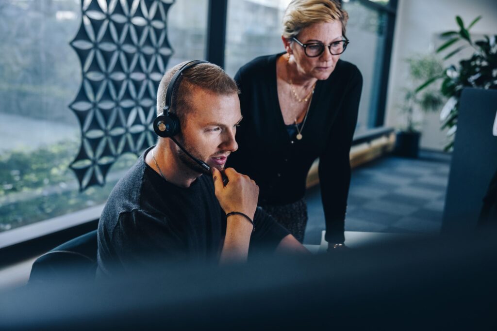 A man wearing a headset sits at a computer while a woman stands beside him, both intently focused on the screen.