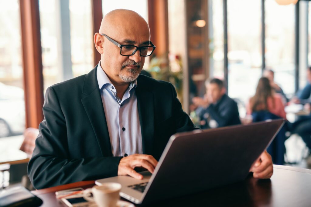 A professional man is seated at a café, typing on his laptop.