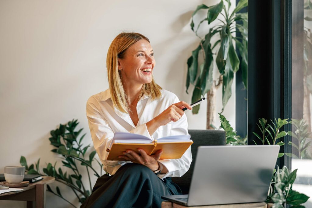 A woman is sitting next to her laptop with an open notebook and a pen in her hands, looking out the window and smiling.