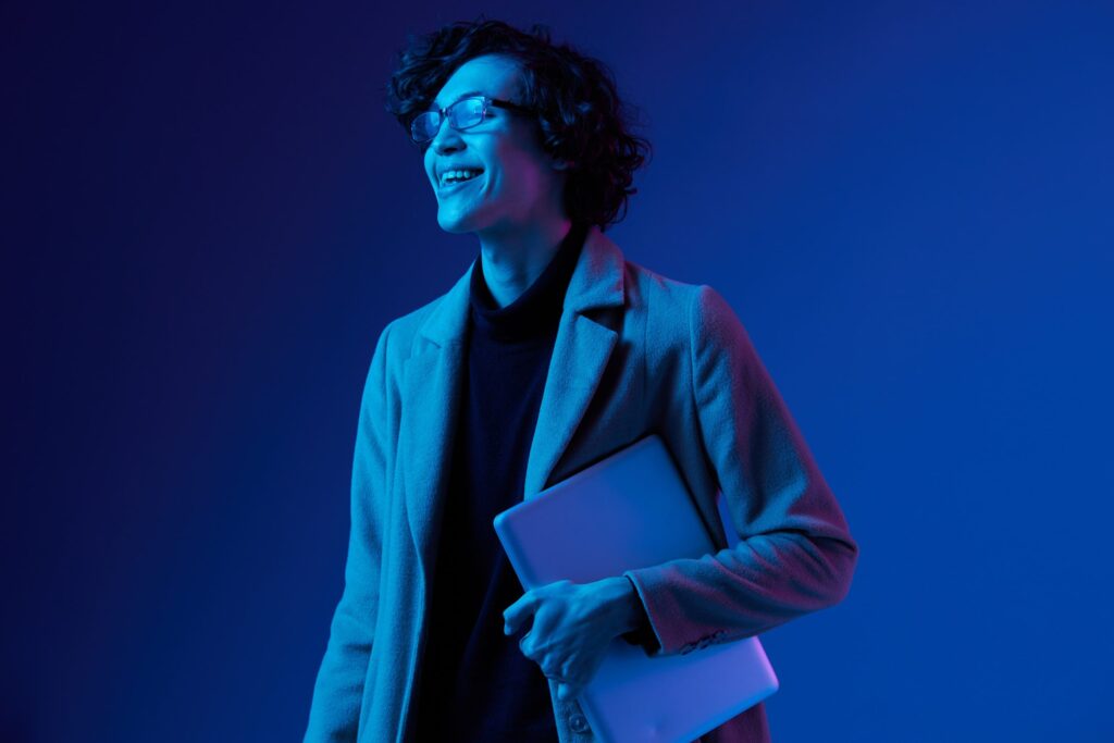 A professionally dressed woman holds a laptop against a blue background.