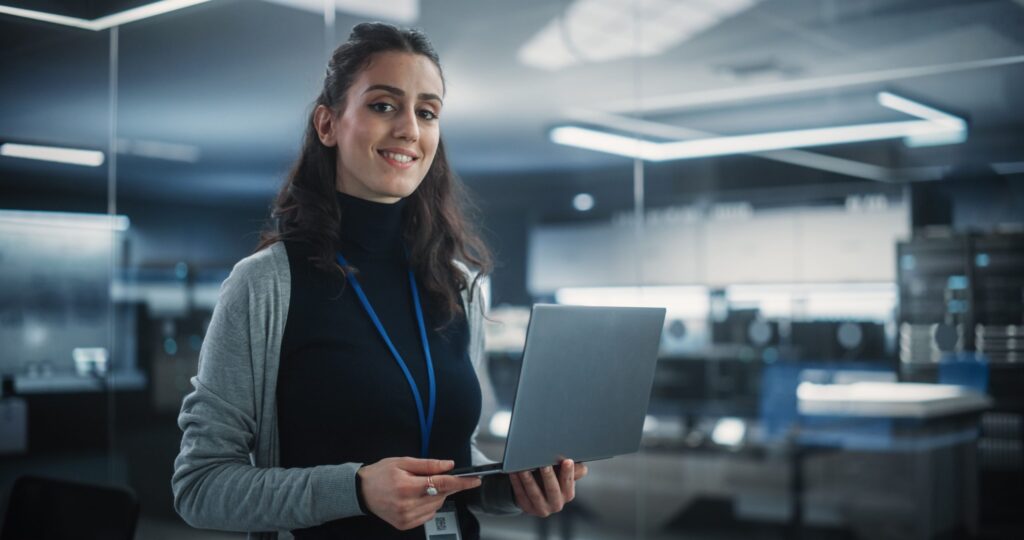a professionally dressed woman is standing in an office setting, holding a laptop. She is looking directly at the camera with a smile.