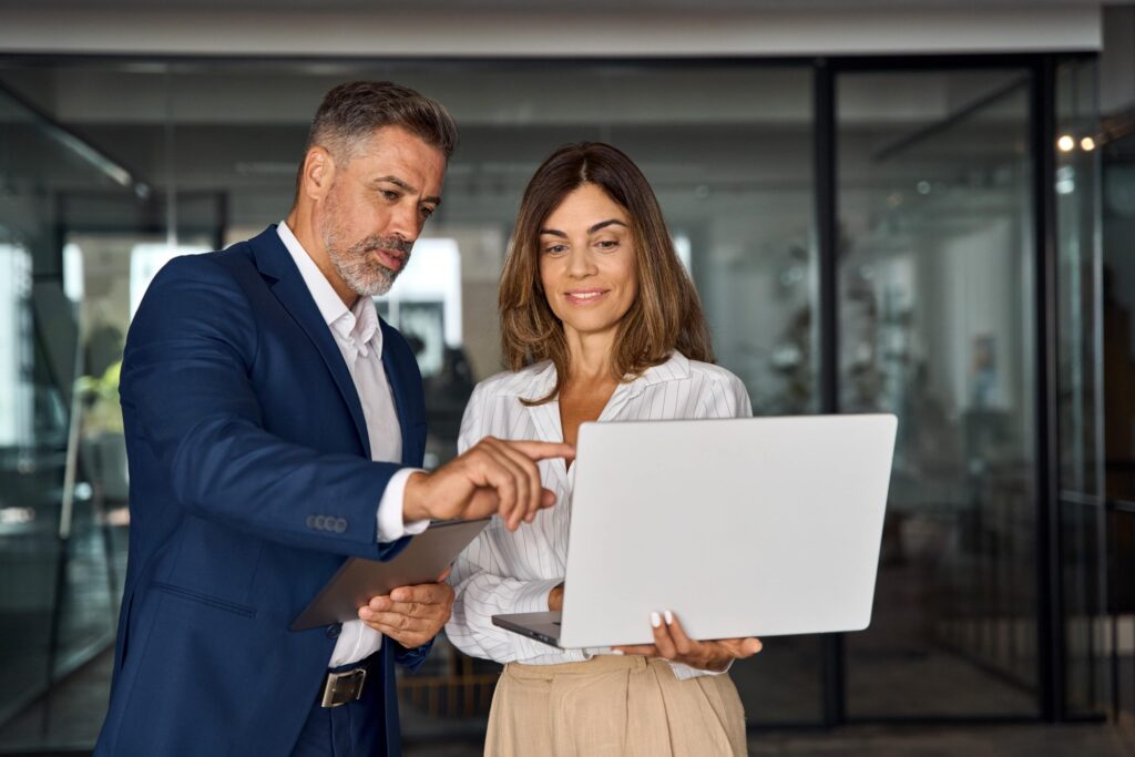 A professionally dressed man and woman are in an office setting, both focused on a laptop screen. The man is pointing at the screen, explaining something, while the woman listens attentively.