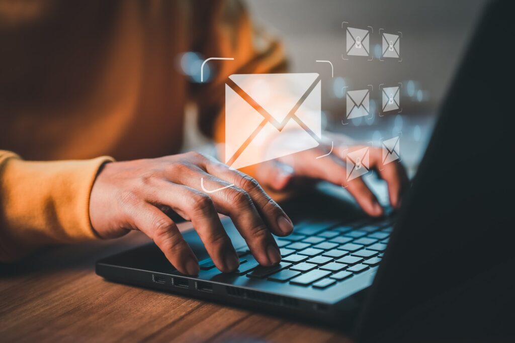 Close-up of a person's hands typing on a laptop keyboard, with a holographic email icon floating above the keyboard and smaller email icons in the background