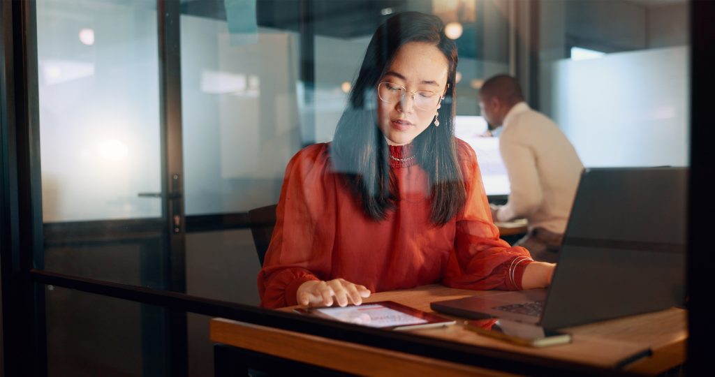 A professionally looking woman in an office, visible through a glass door, working on an open laptop while looking at a tablet and typing.